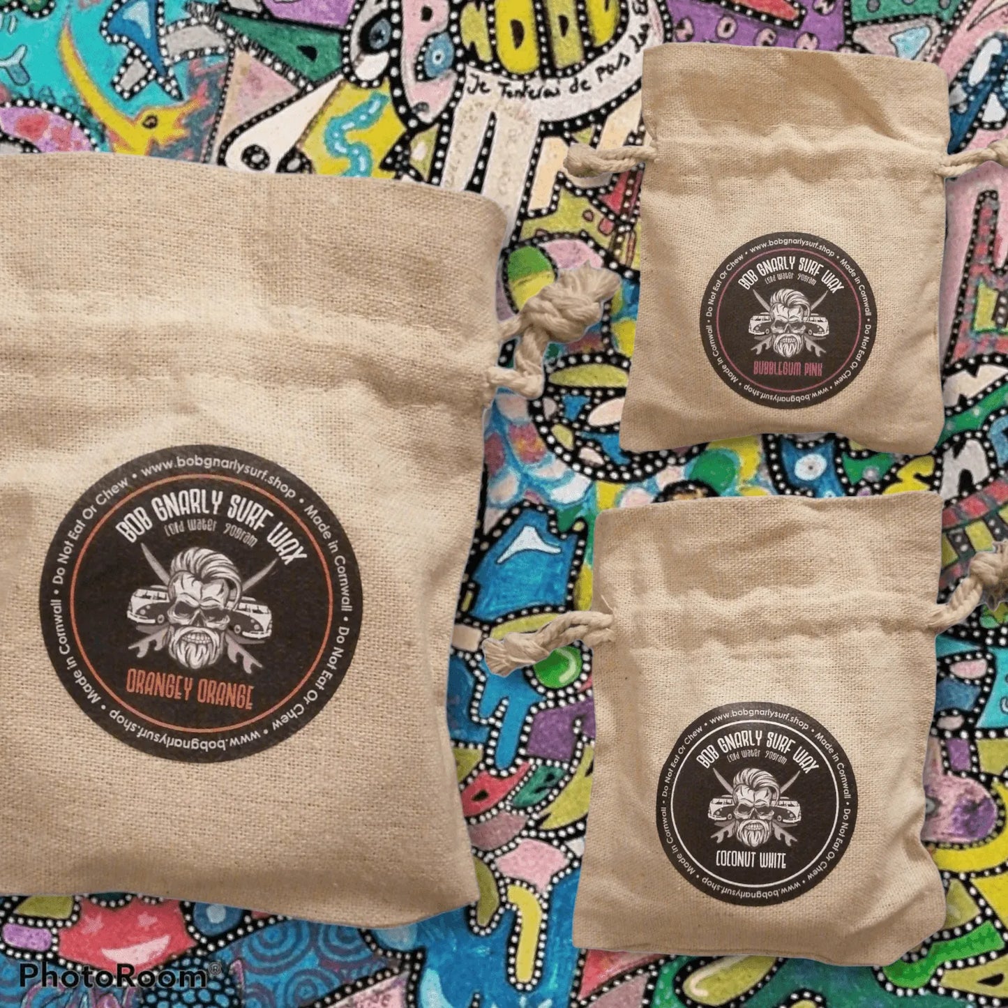 Three beige drawstring bags of Bob Gnarly Surf Wax on colorful patterned background, labeled Orangey Orange, Bubblegum Pink, Coconut White