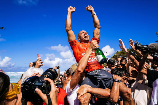 Surfer wearing red shirt lifted by crowd holding champagne at beach with blue sky