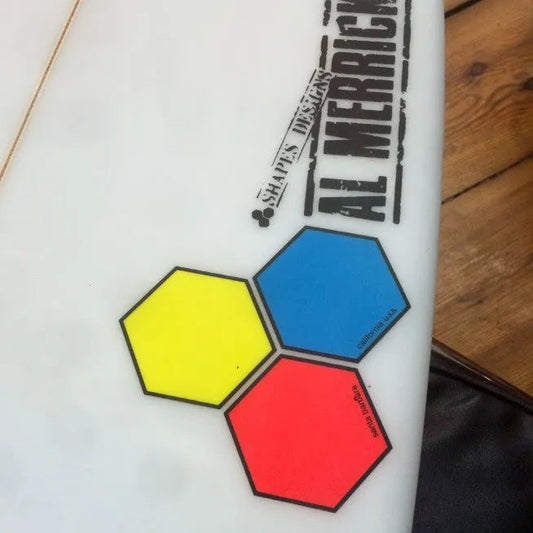 Close-up of white Al Merrick surfboard with colorful hexagon logos in yellow, blue, and red on wooden floor