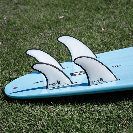 Light blue surfboard with four white FCS II fins on green grass background