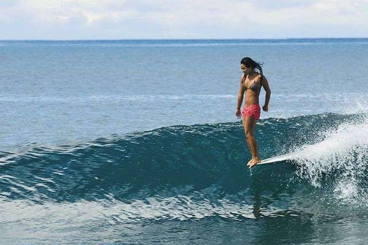 Woman surfing on a clear blue ocean wave wearing pink shorts and a bikini top