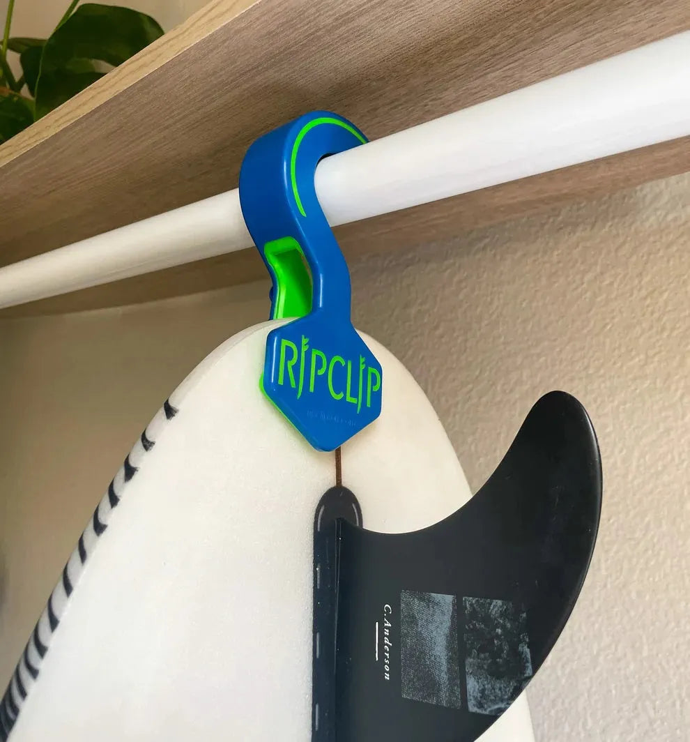 Blue and green RipClip hanger holding a white surfboard with a black fin on a white rod under a wooden shelf