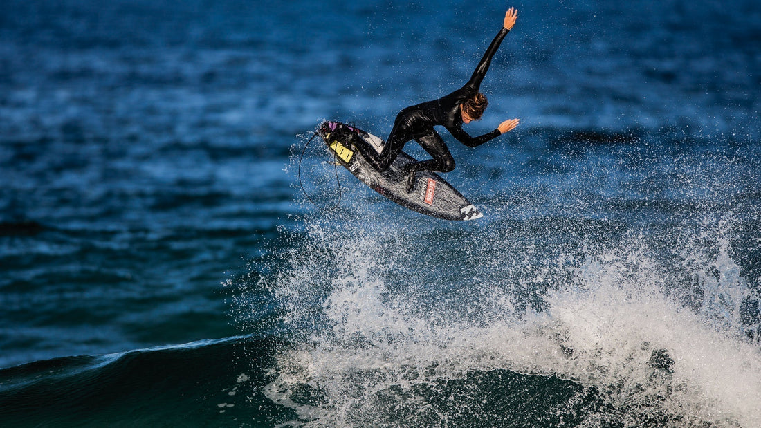 Surfer in black wetsuit performing aerial trick above ocean wave with surfboard splashing water