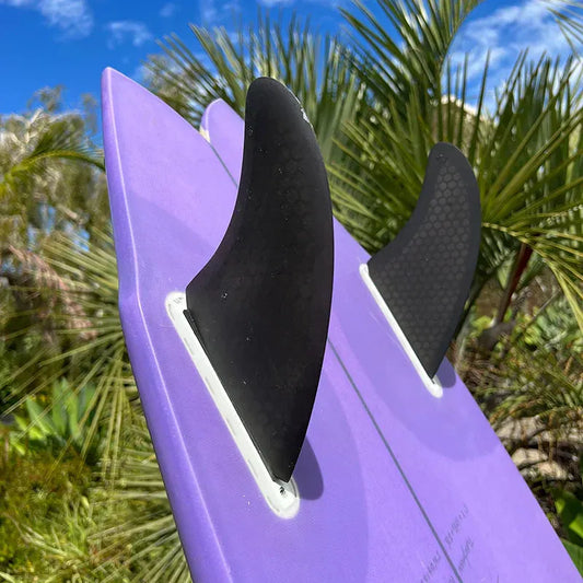 Close-up of purple twin-fin surfboard with black fins against green palm trees and blue sky