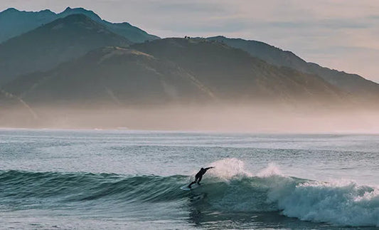Surfer riding a wave in the ocean with misty mountains in the background at sunrise