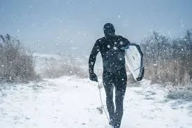 Person in black wetsuit carrying surfboard walking through snowy, windy landscape