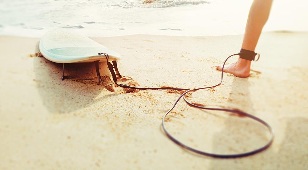 Surfboard leash attached to barefoot person on sandy beach near ocean water
