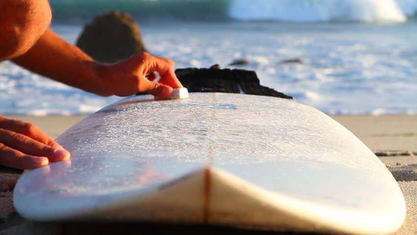 Close-up of hands waxing a white surfboard on sandy beach with ocean waves in the background