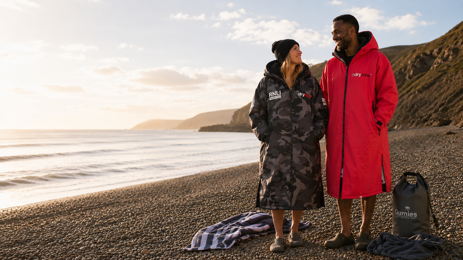 Person wearing a black and pink Dryrobe holding a surfboard outdoors with rocky background