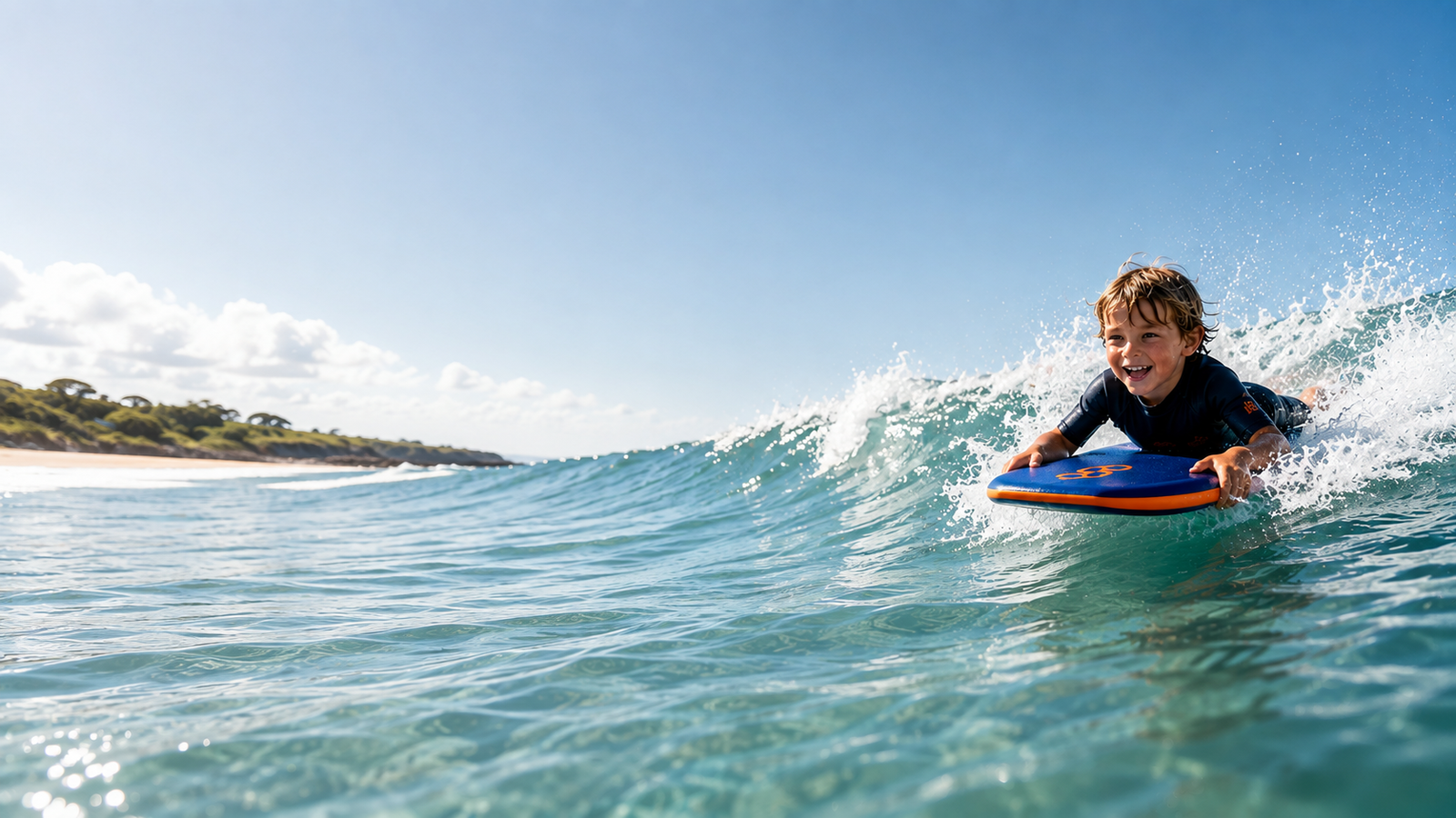 Smiling boy in yellow wetsuit holding colorful bodyboard on sandy beach with ocean and green cliffs