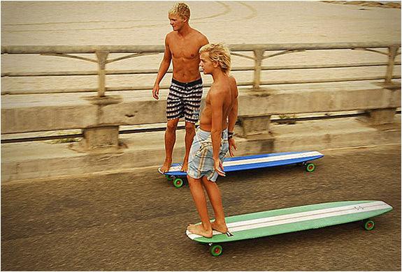 Two shirtless boys skateboarding on longboards by a seaside concrete railing under bright daylight