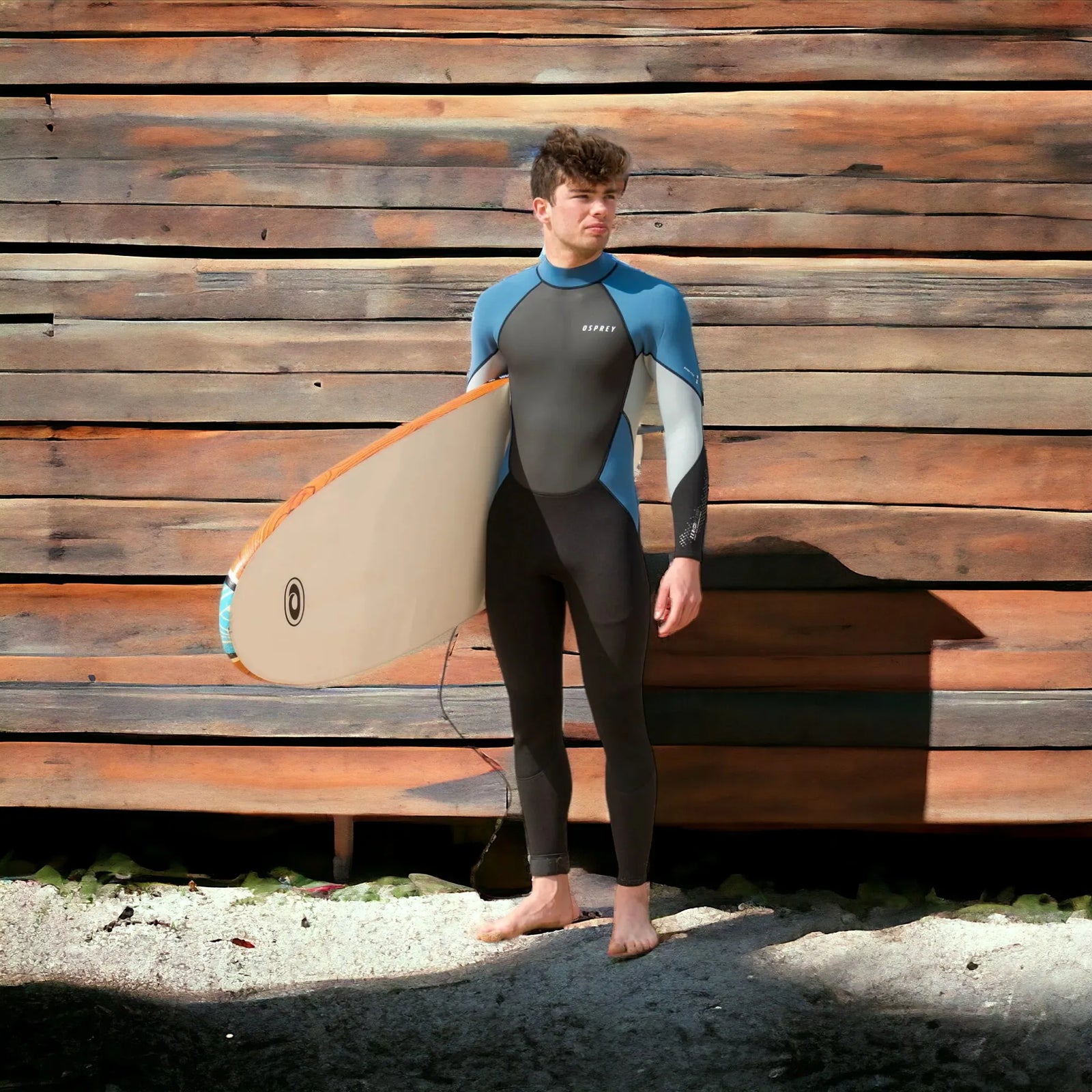 Man in Osprey wetsuit holding surfboard, standing barefoot on sand in front of wooden wall