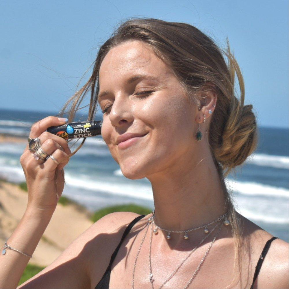 Young woman holding SPF 50 zinc stick sunscreen near face at sunny beach with ocean background