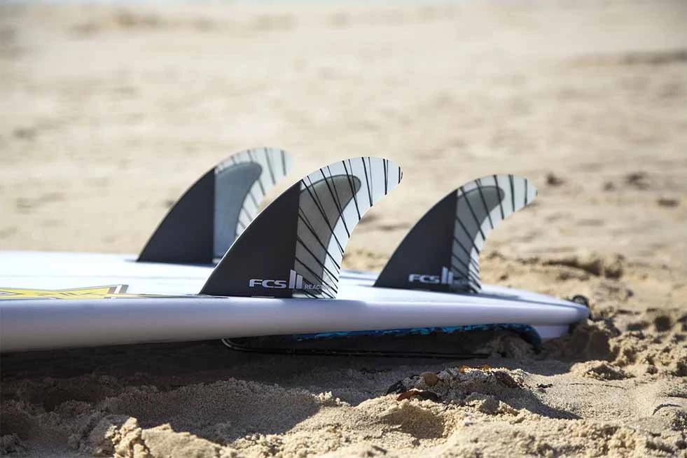 White surfboard with three black and white FCS II fins resting on sandy beach